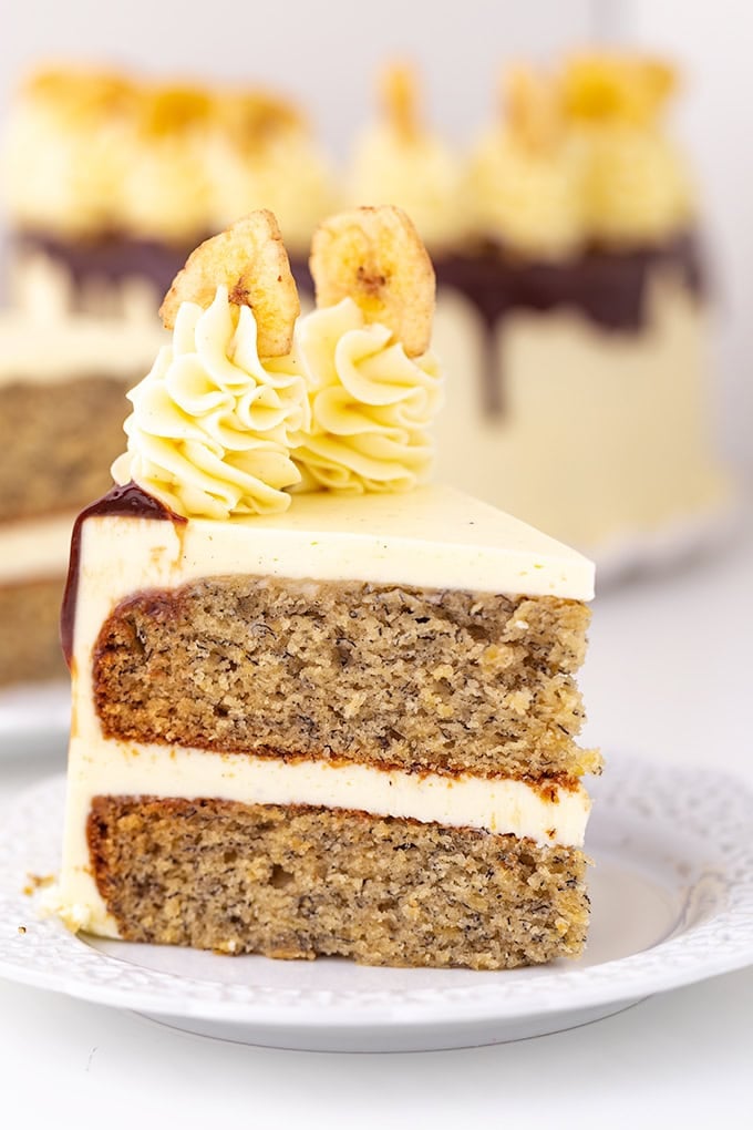 A slice of Roasted Banana Cream Cake with light frosting, chocolate drizzle, and piped cream topped with banana chips, served on a white plate.