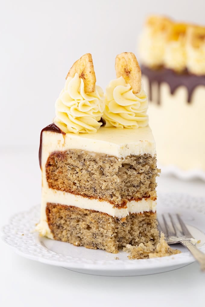A slice of Roasted Banana Cream Cake with vanilla frosting, two banana chip decorations on top, and a fork on a white plate.