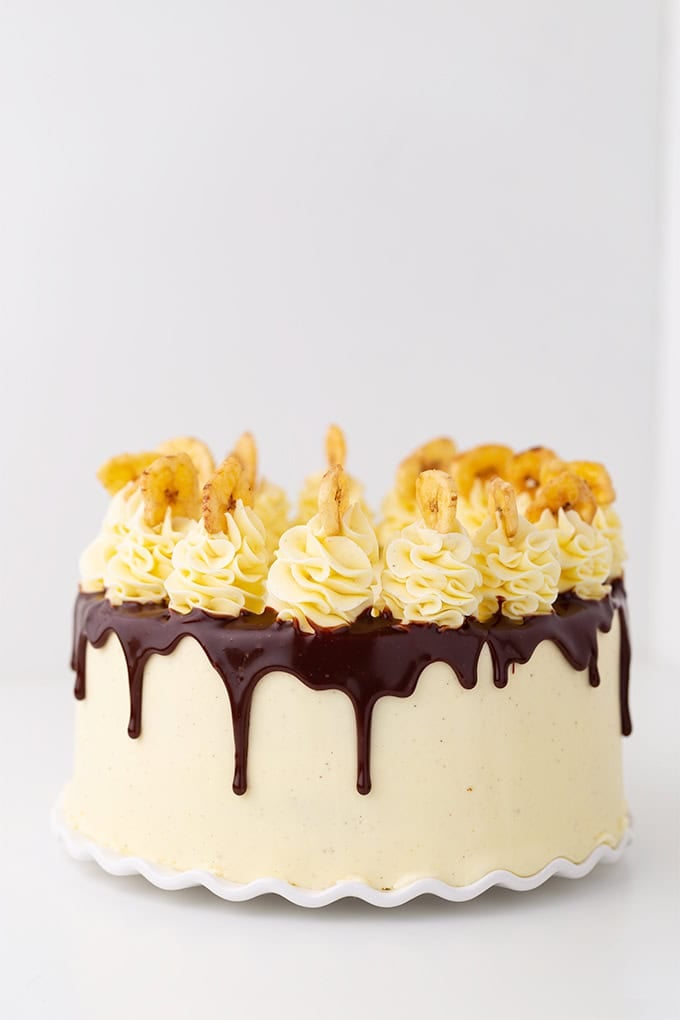 A round Roasted Banana Cream Cake with vanilla frosting, chocolate ganache dripping over the sides, piped cream on top, and pieces of dried banana as decoration.