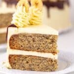 A slice of Roasted Banana Cream Cake with two layers of frosting, chocolate drizzle, and piped cream topped with banana chips, served on a white plate.