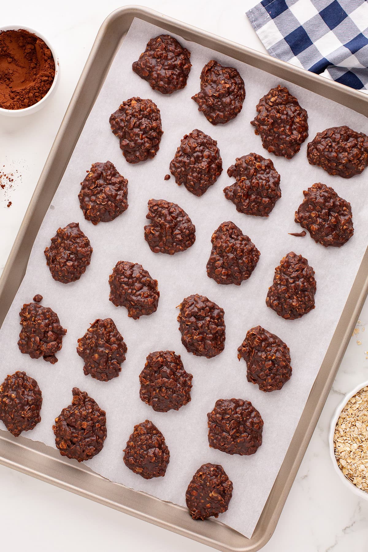 A baking sheet lined with parchment paper holds several Chocolate No-Bake Cookies, with bowls of cocoa powder and oats nearby.