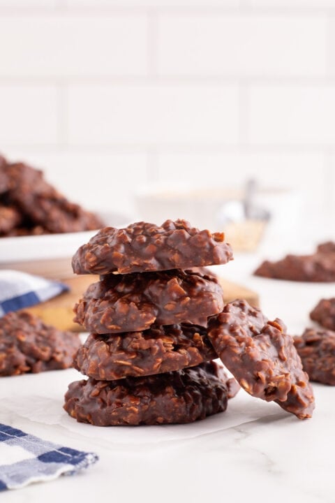 A stack of five Chocolate No-Bake Cookies sits on a white surface, with more cookies and ingredients softly blurred in the background.