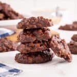 A stack of Chocolate No-Bake Cookies with oats sits on parchment paper, surrounded by more cookies on a white surface.