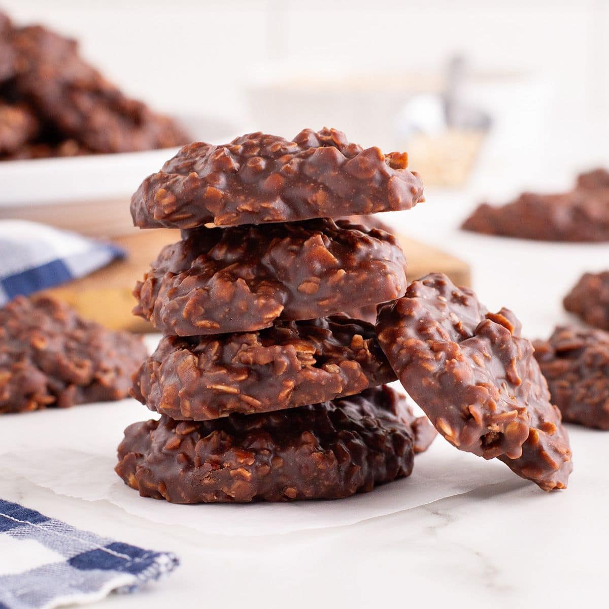 A stack of Chocolate No-Bake Cookies with oats sits on parchment paper, surrounded by more cookies on a white surface.