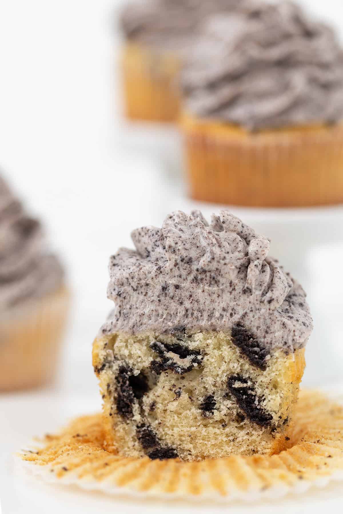 A partially eaten Oreo Cookies and Cream Cupcake with cookies and cream frosting sits on an unwrapped liner, revealing chunks of cookie inside. More frosted cupcakes are visible in the background.