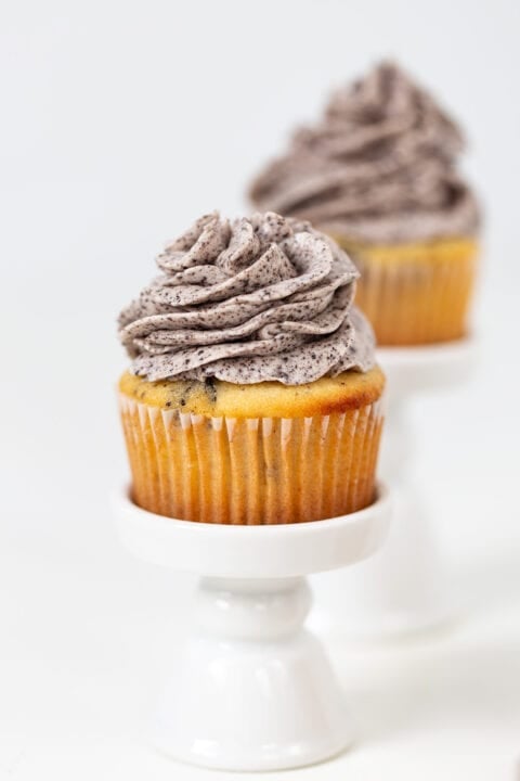 An Oreo Cookies and Cream Cupcake with vanilla base and cookies and cream frosting is displayed on a small white stand, with another cupcake blurred in the background.