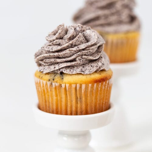 An Oreo Cookies and Cream Cupcake with vanilla base and cookies and cream frosting is displayed on a small white stand, with another cupcake blurred in the background.
