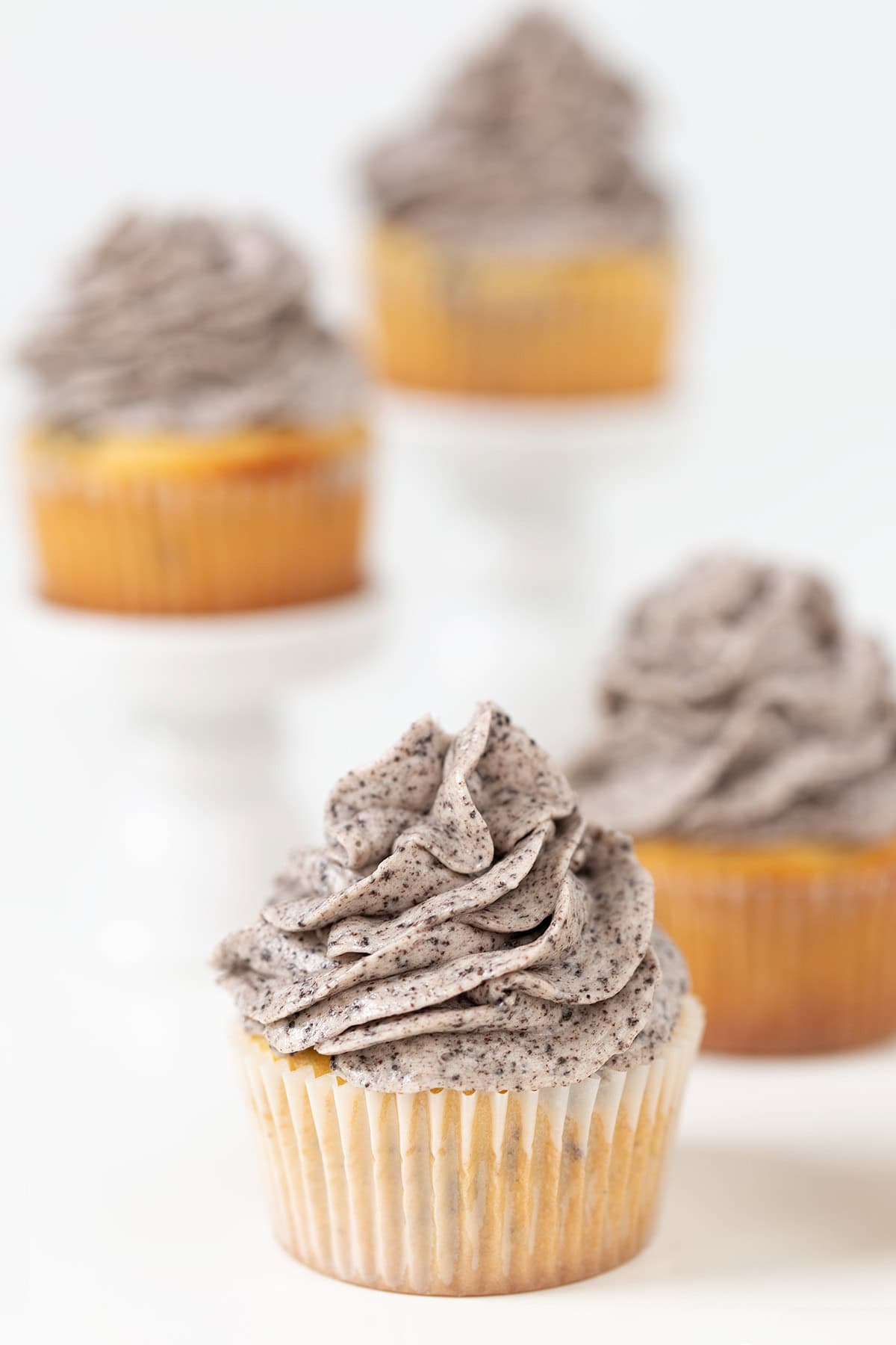 Four Oreo Cookies and Cream Cupcakes with rich cookies and cream frosting, displayed on a white surface, with two elevated on white stands in the background.