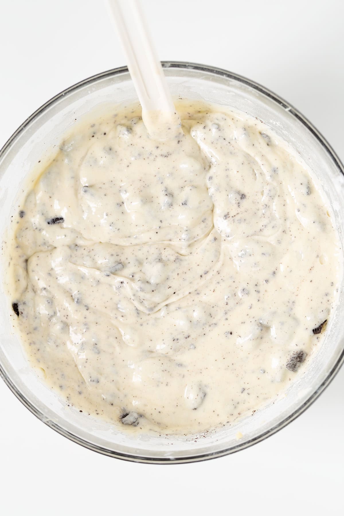 A glass bowl filled with a creamy, speckled Oreo Cookies and Cream Cupcakes batter is being mixed with a spatula.