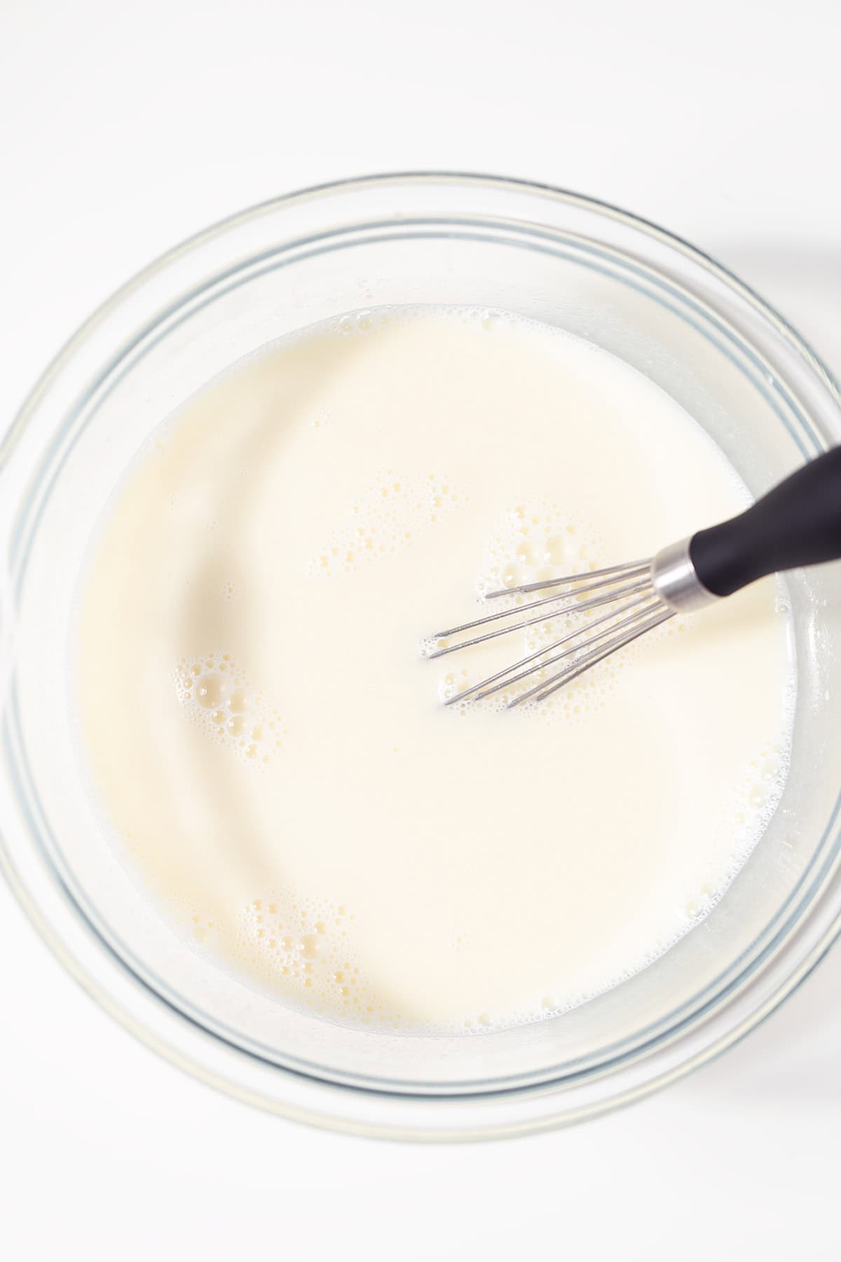 A glass bowl filled with a creamy mixture is being whisked on a white surface, perfect for creating the luscious base of Oreo Cookies and Cream Cupcakes.