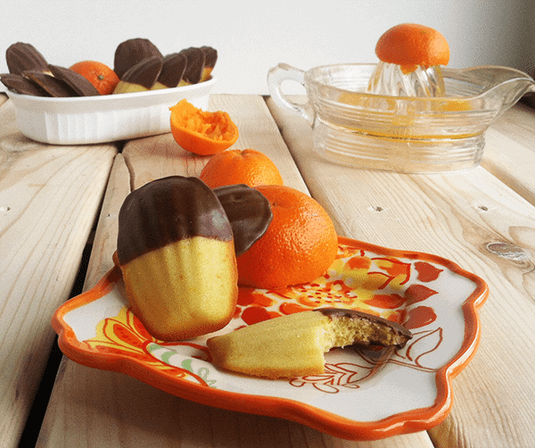 A plate of chocolate covered oranges with orange madeleines on a wooden table.