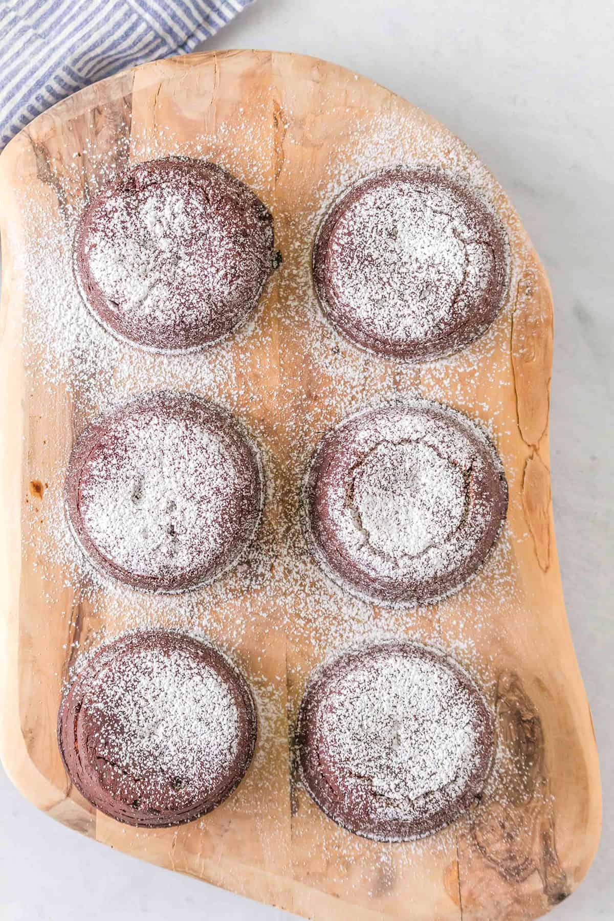 Six decadent Chocolate Lava Cakes dusted with powdered sugar, elegantly arranged on a wooden cutting board.