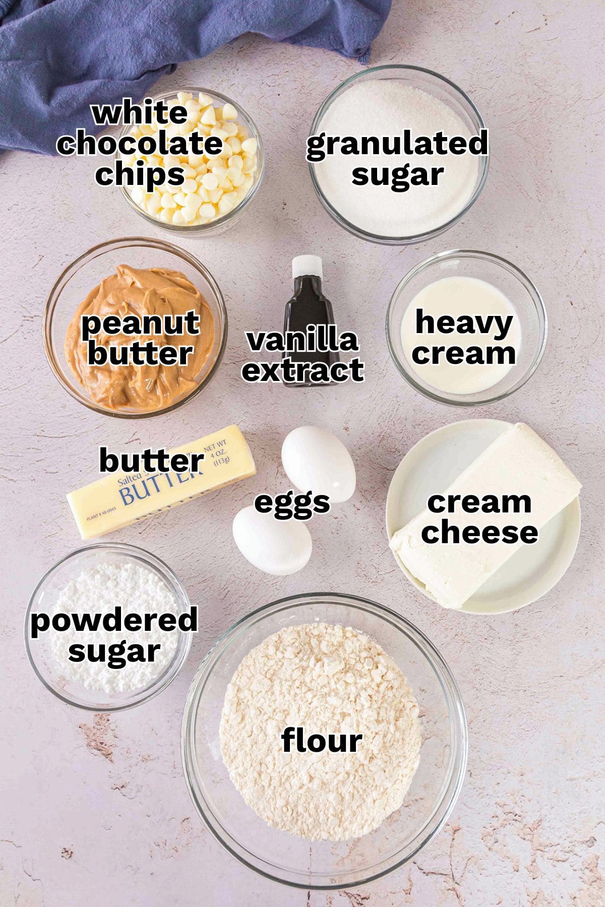 Top-down view of ingredients for white chocolate brownies with peanut butter frosting: white chocolate chips, granulated sugar, peanut butter, vanilla extract, heavy cream, butter, eggs, cream cheese, powdered sugar, and flour.