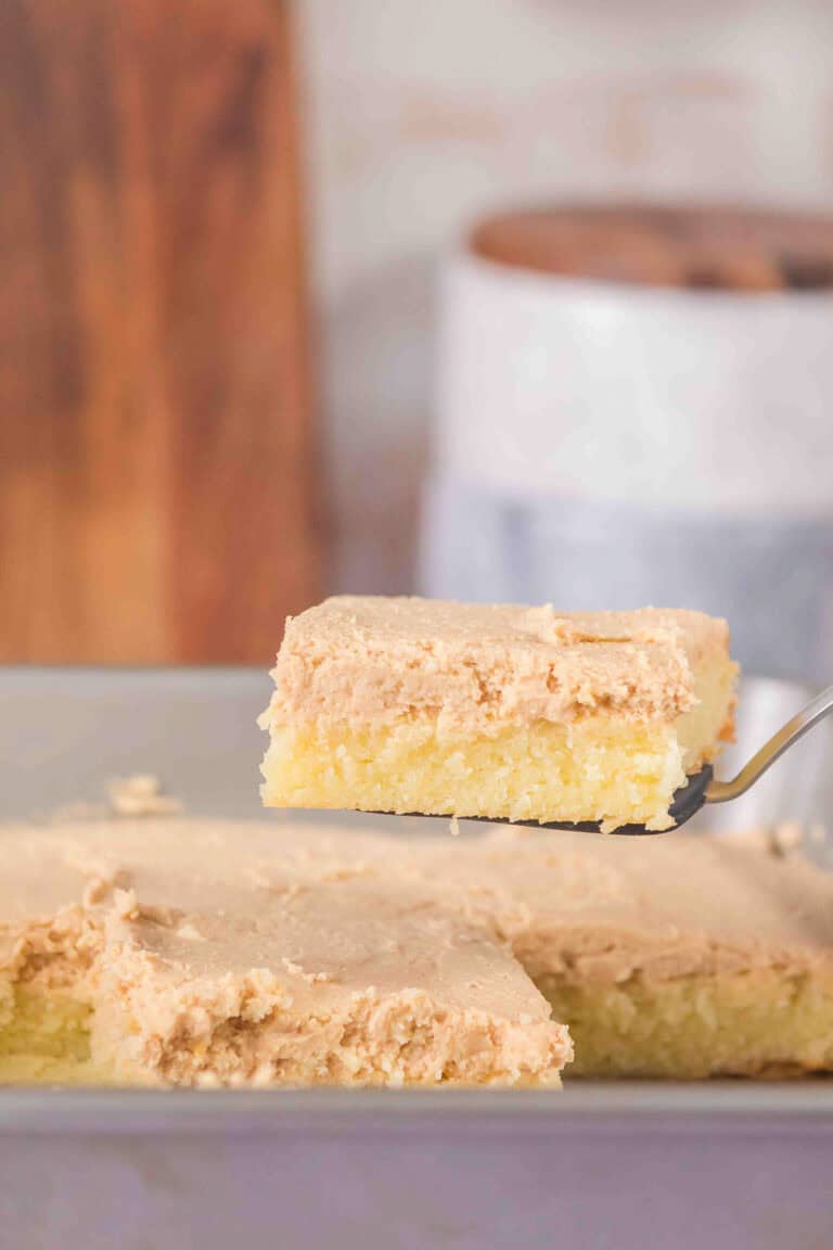 A close-up of a slice of white chocolate brownies with peanut butter frosting being lifted from a baking dish with a spatula.