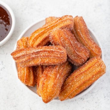 A plate of sugar-coated churros is displayed on a white surface, with a small bowl of chocolate sauce partially visible in the corner, inviting you to dip each churro into the rich, velvety treat.