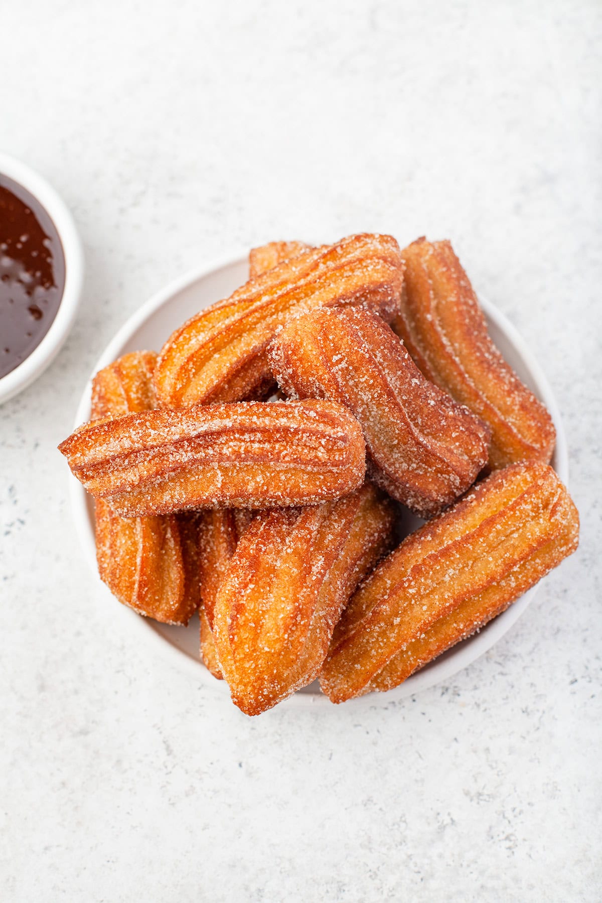 A plate of sugar-coated churros is displayed on a white surface, with a small bowl of chocolate sauce partially visible in the corner, inviting you to dip each churro into the rich, velvety treat.