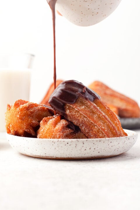 Golden churros are being drizzled with rich chocolate sauce, accompanied by a glass of milk and extra churros in the background.