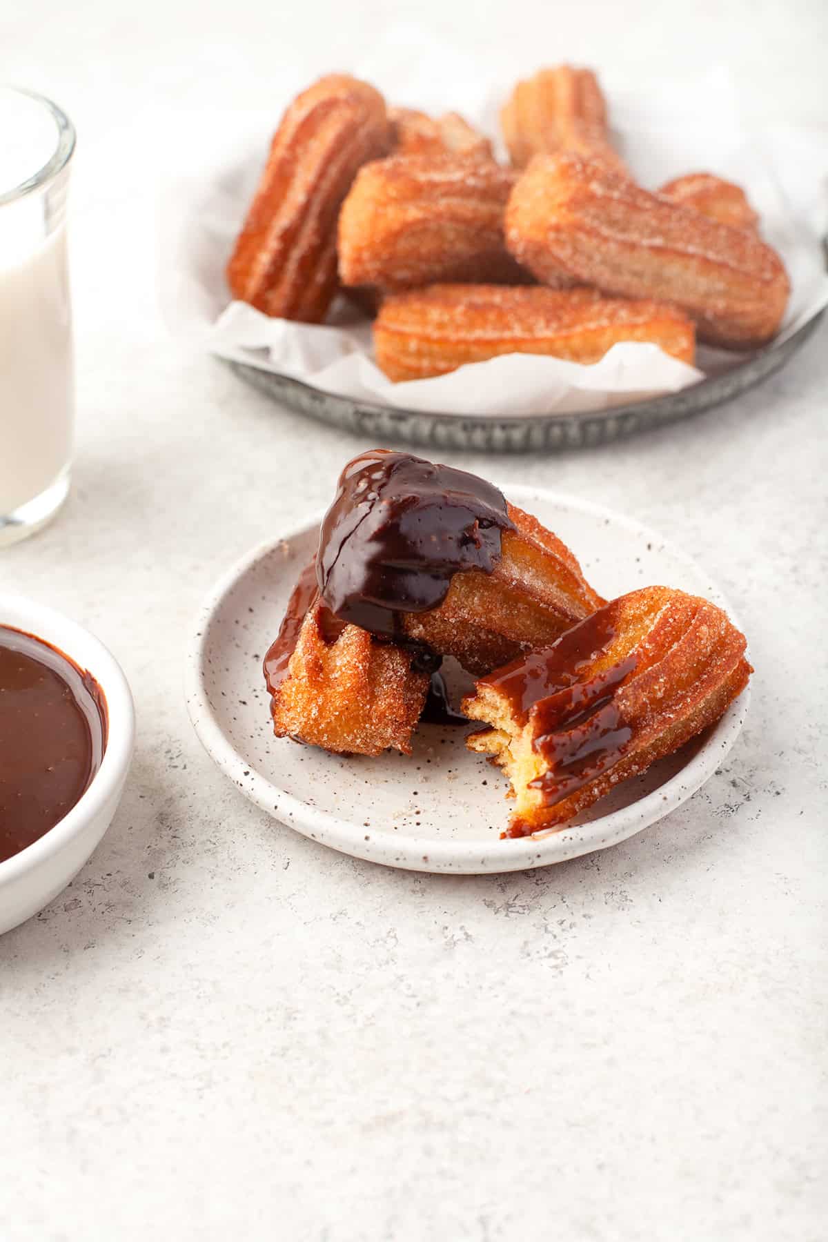 A small plate with churros drizzled in chocolate sauce, a bowl of rich chocolate sauce, a glass of milk, and an extra plate of plain churros in the background create a tempting churros spread.
