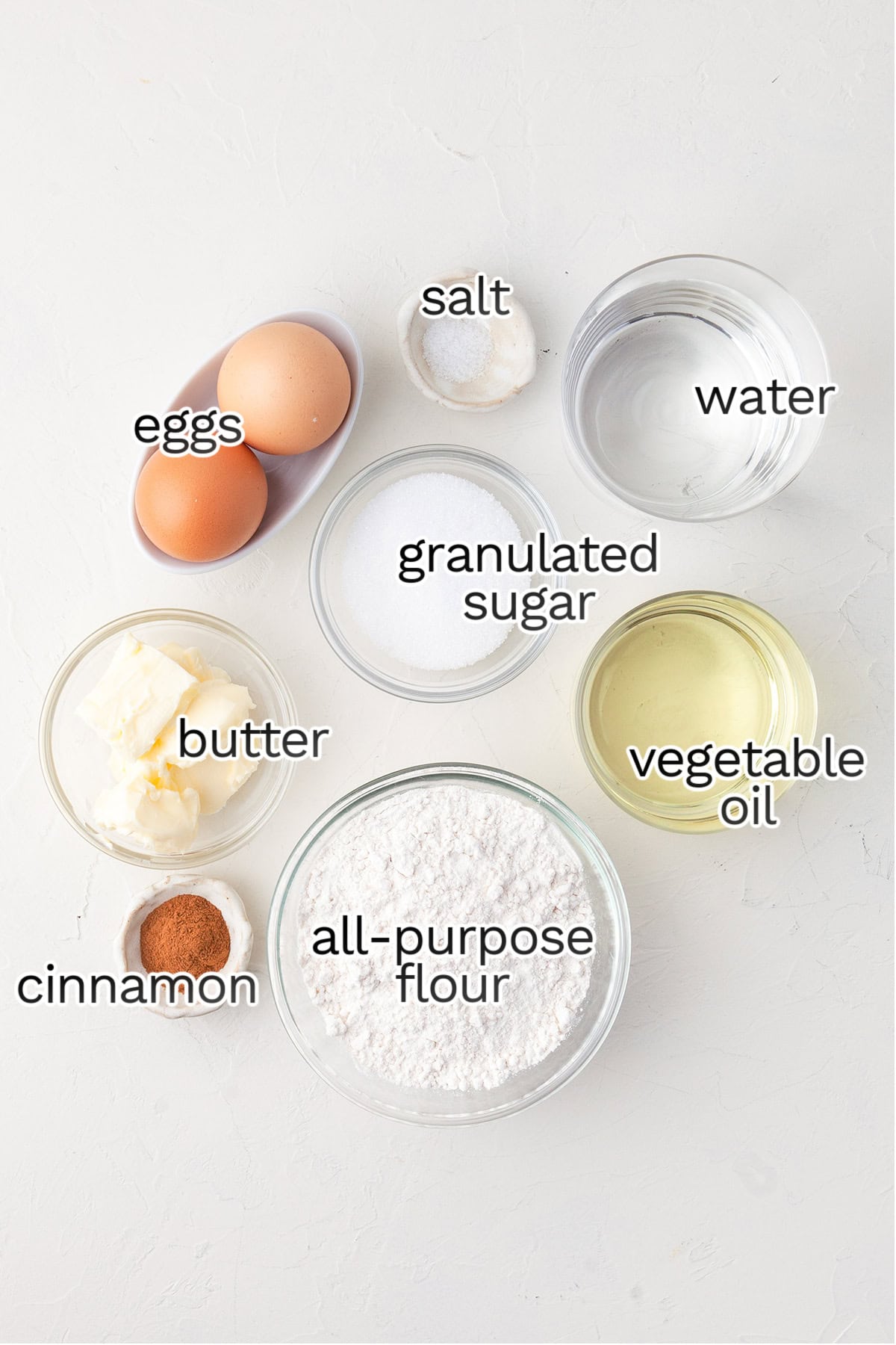 Measured ingredients for homemade churros are arranged in separate bowls on a white surface, including eggs, salt, water, sugar, vegetable oil, butter, cinnamon, and all-purpose flour.