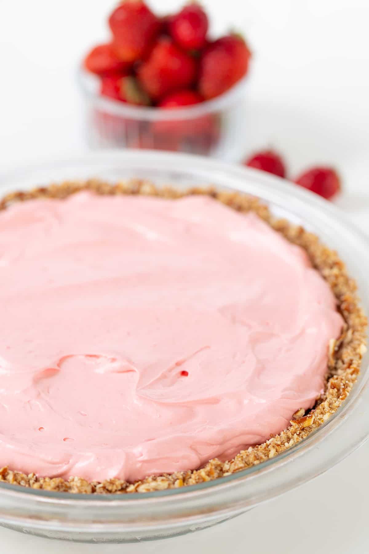 A Strawberry Cream Pie with a pink creamy filling sits in a glass dish with a crumbly crust. A small bowl of strawberries is in the background.