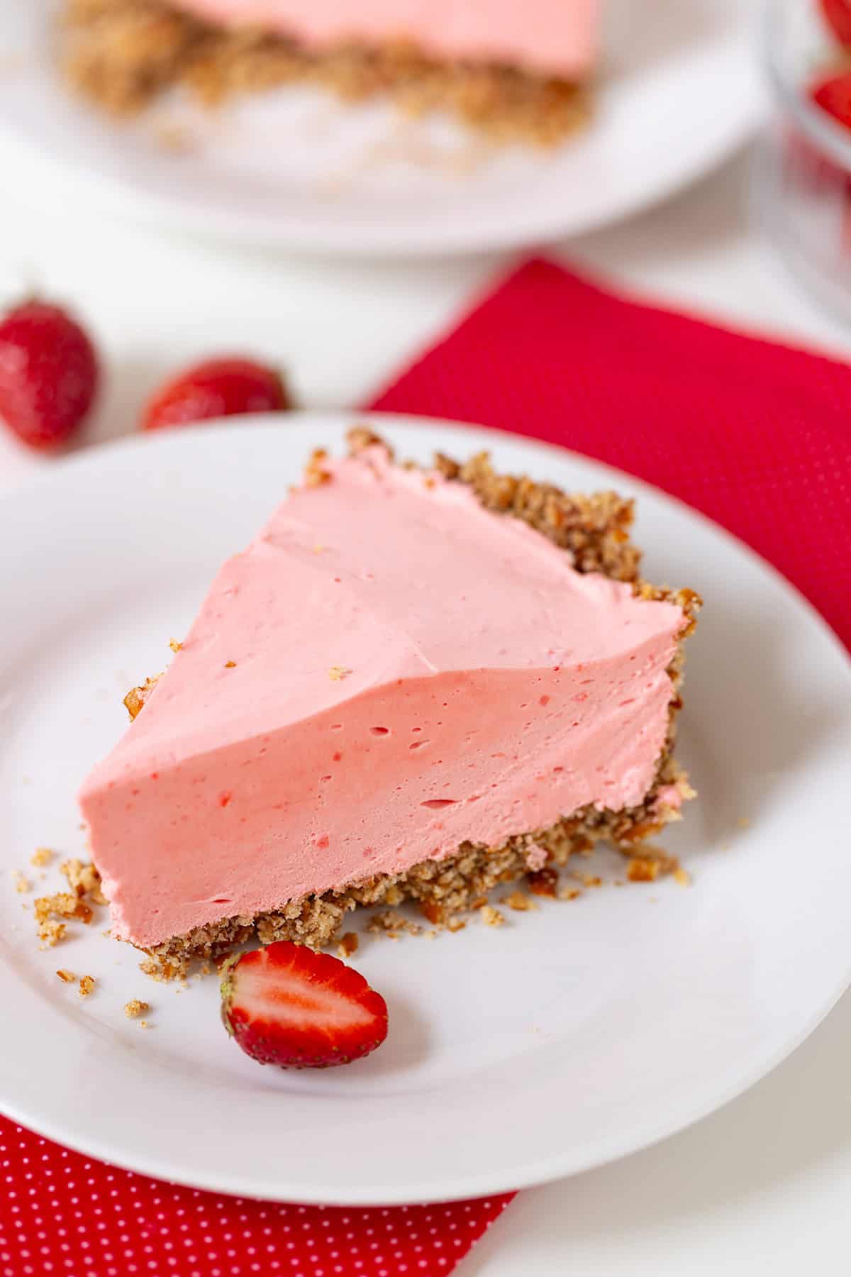 A slice of pink strawberry cream pie graces a white plate, showcasing its rich graham cracker crust and a fresh sliced strawberry garnish. Beneath it lies a vibrant red fabric napkin, adding an extra touch of elegance.