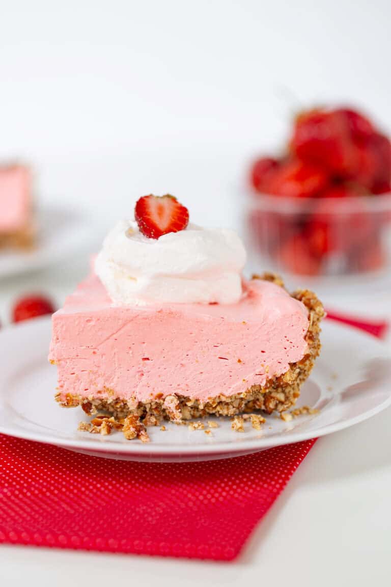 A delectable slice of pink strawberry cream pie on a white plate features a graham cracker crust, elegantly topped with whipped cream and a fresh strawberry slice. In the background, a bowl of strawberries adds to the charm.