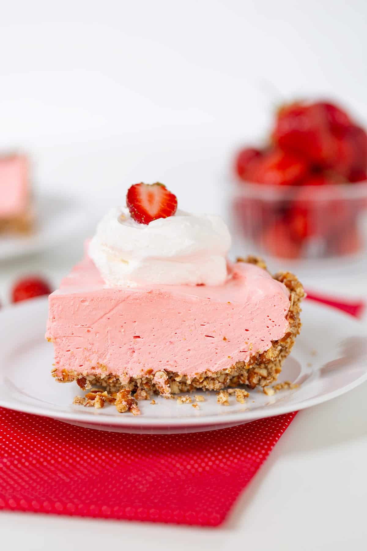 A delectable slice of pink strawberry cream pie on a white plate features a graham cracker crust, elegantly topped with whipped cream and a fresh strawberry slice. In the background, a bowl of strawberries adds to the charm.