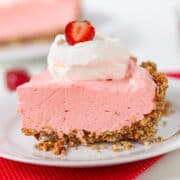 A slice of strawberry cream pie with a crumb crust, topped with whipped cream and a strawberry slice, rests elegantly on a white plate with a red napkin.