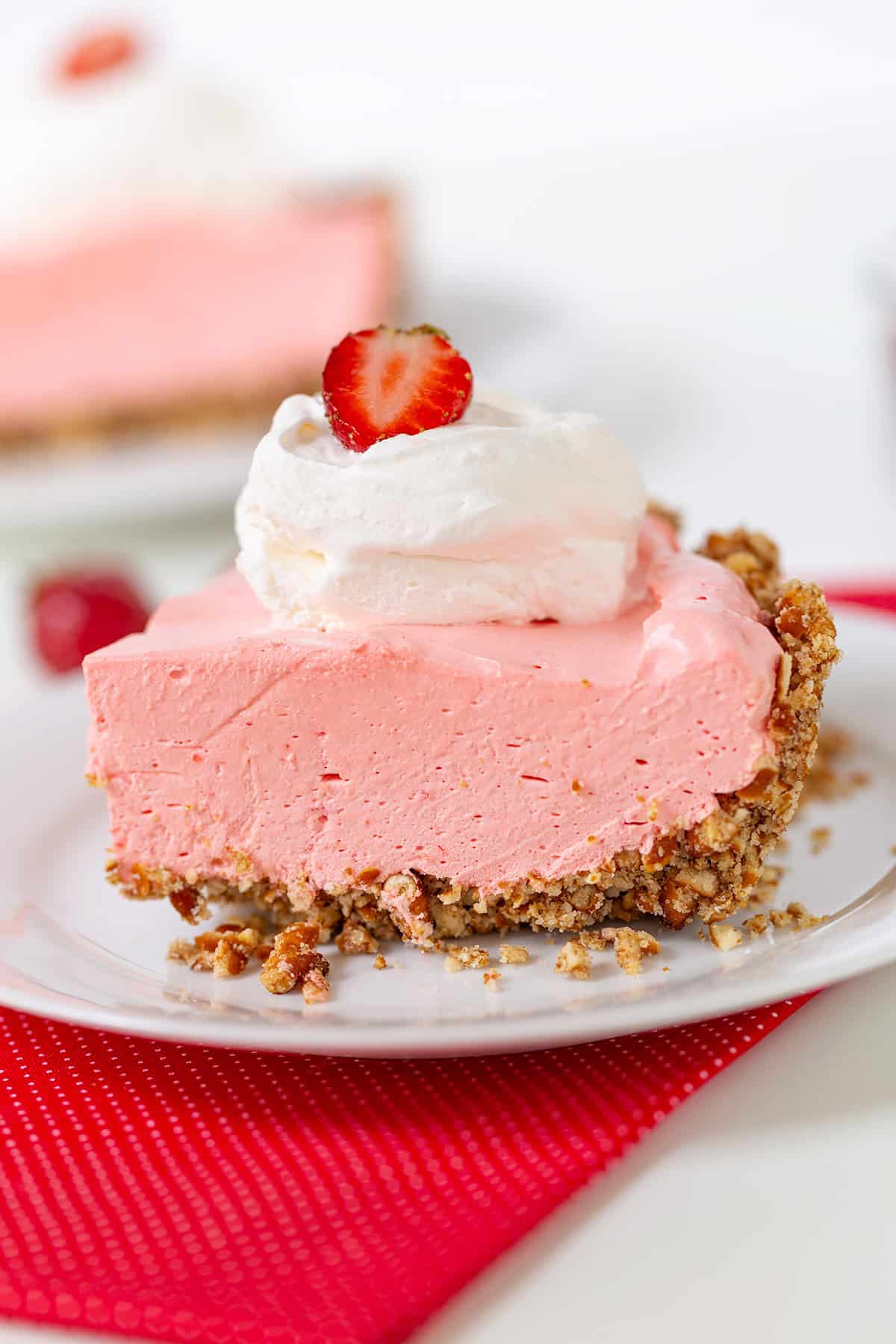 A slice of strawberry cream pie with a crumb crust, topped with whipped cream and a strawberry slice, rests elegantly on a white plate with a red napkin.