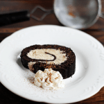 A slice of Chocolate Rolled Cake with Cookie Dough Buttercream and a dollop of whipped cream sits on a white plate, set on a wooden table.