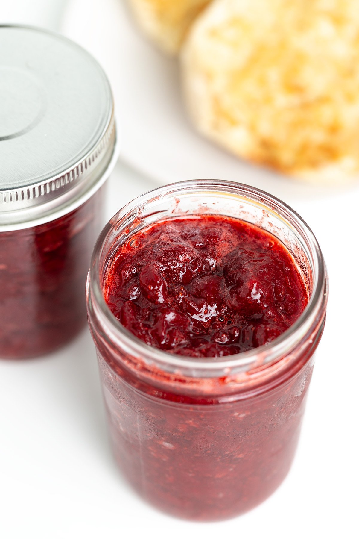 A glass jar filled with strawberry jam sits next to another jar and a plate with toasted bread in the background.