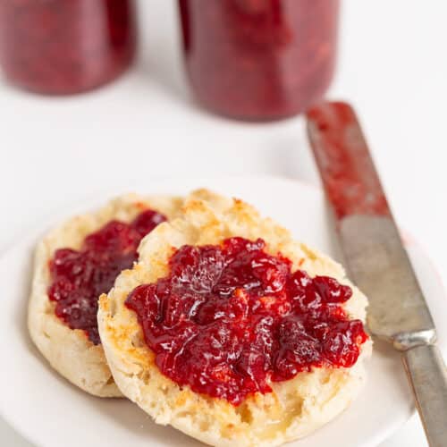 An English muffin split in half with strawberry jam spread on top, placed on a white plate with a jam-covered knife beside it; jars of jam are in the background.