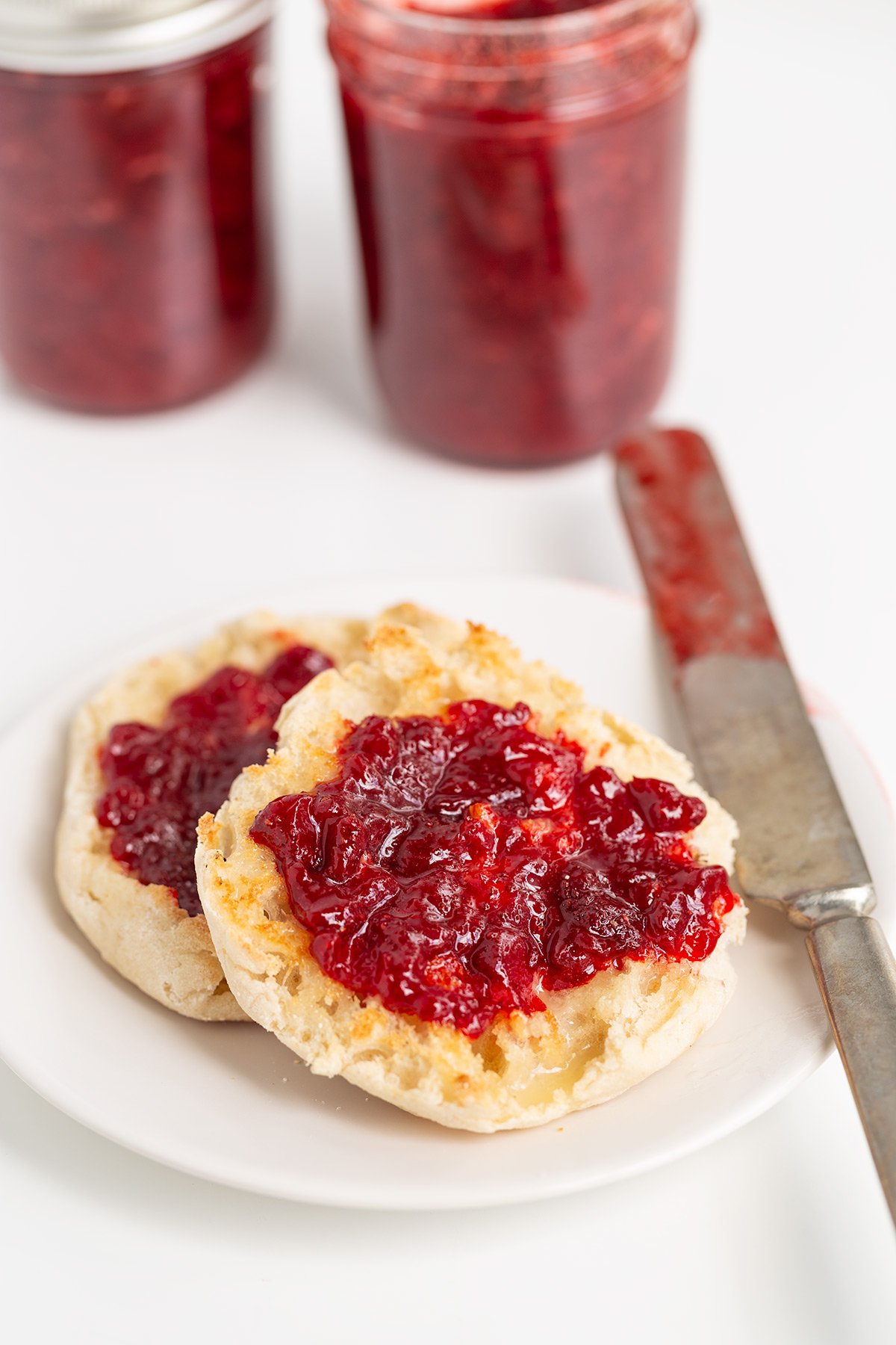 An English muffin split in half with strawberry jam spread on top, placed on a white plate with a jam-covered knife beside it; jars of jam are in the background.