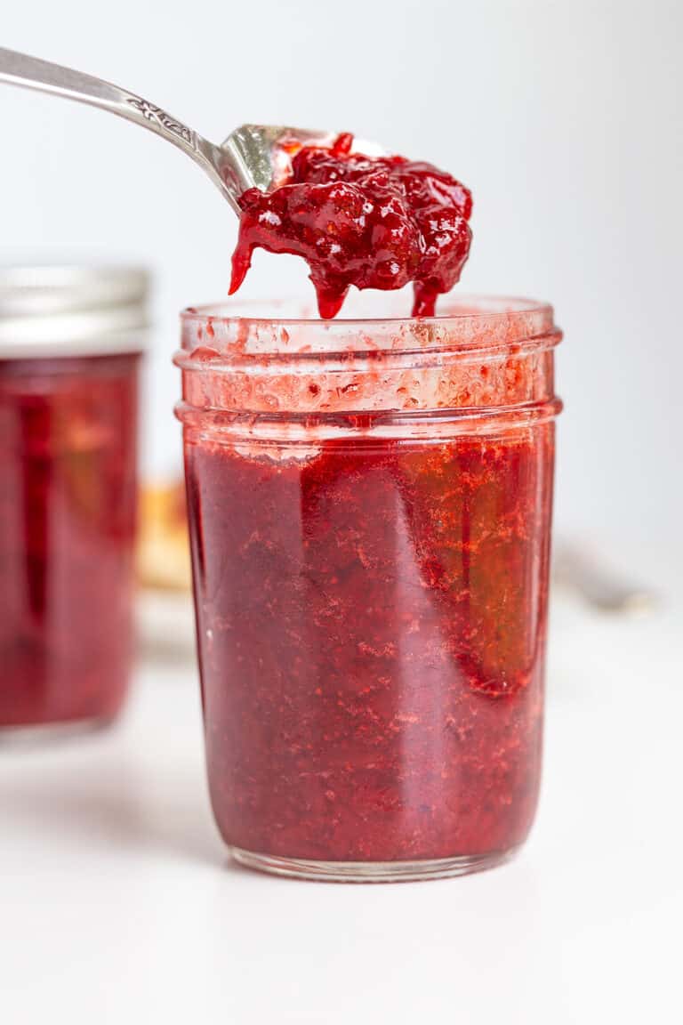 A spoon holding a portion of strawberry jam is held above a glass jar filled with the same sweet jam, with another jar in the background.