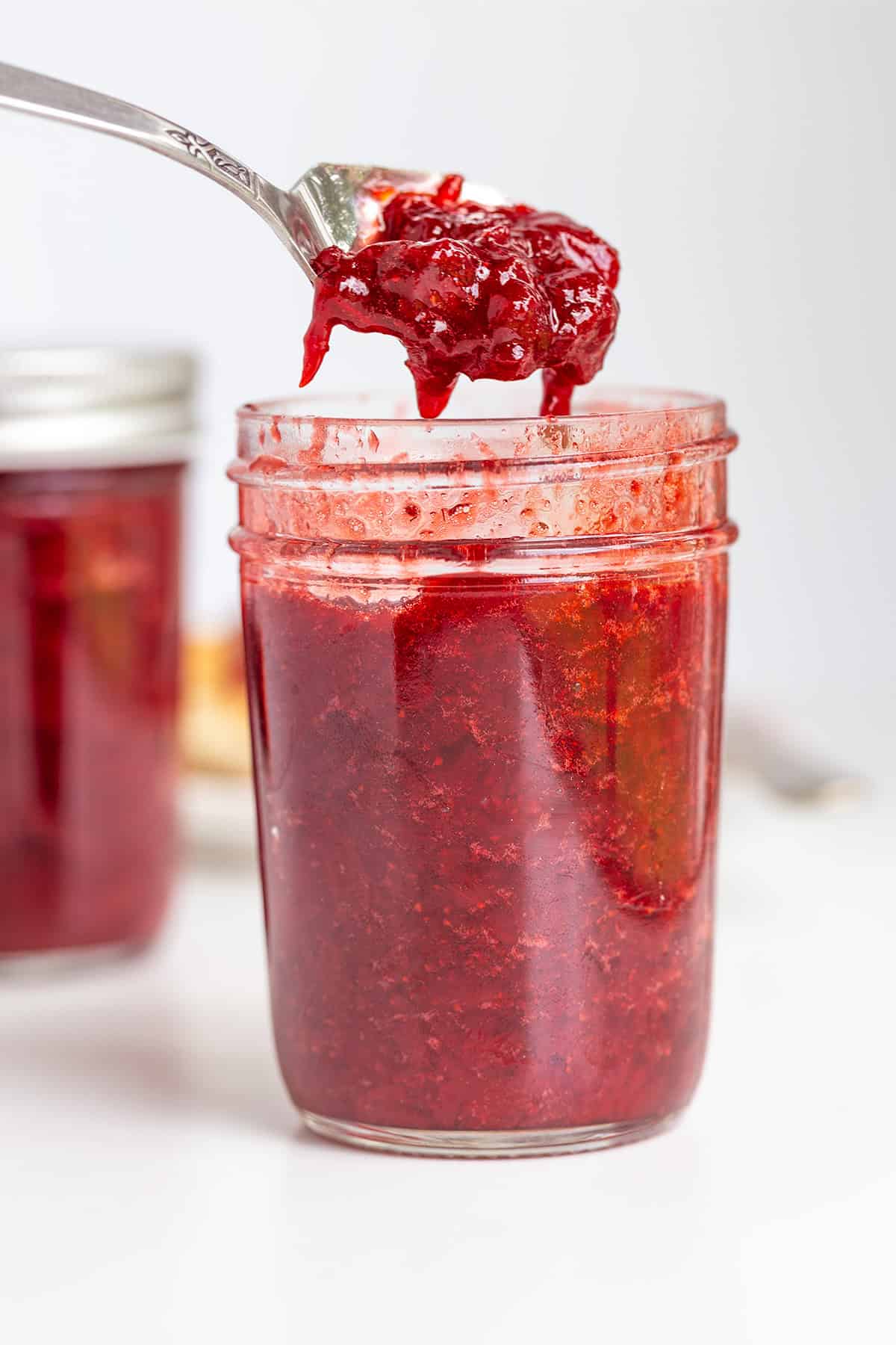 A spoon holding a portion of strawberry jam is held above a glass jar filled with the same sweet jam, with another jar in the background.