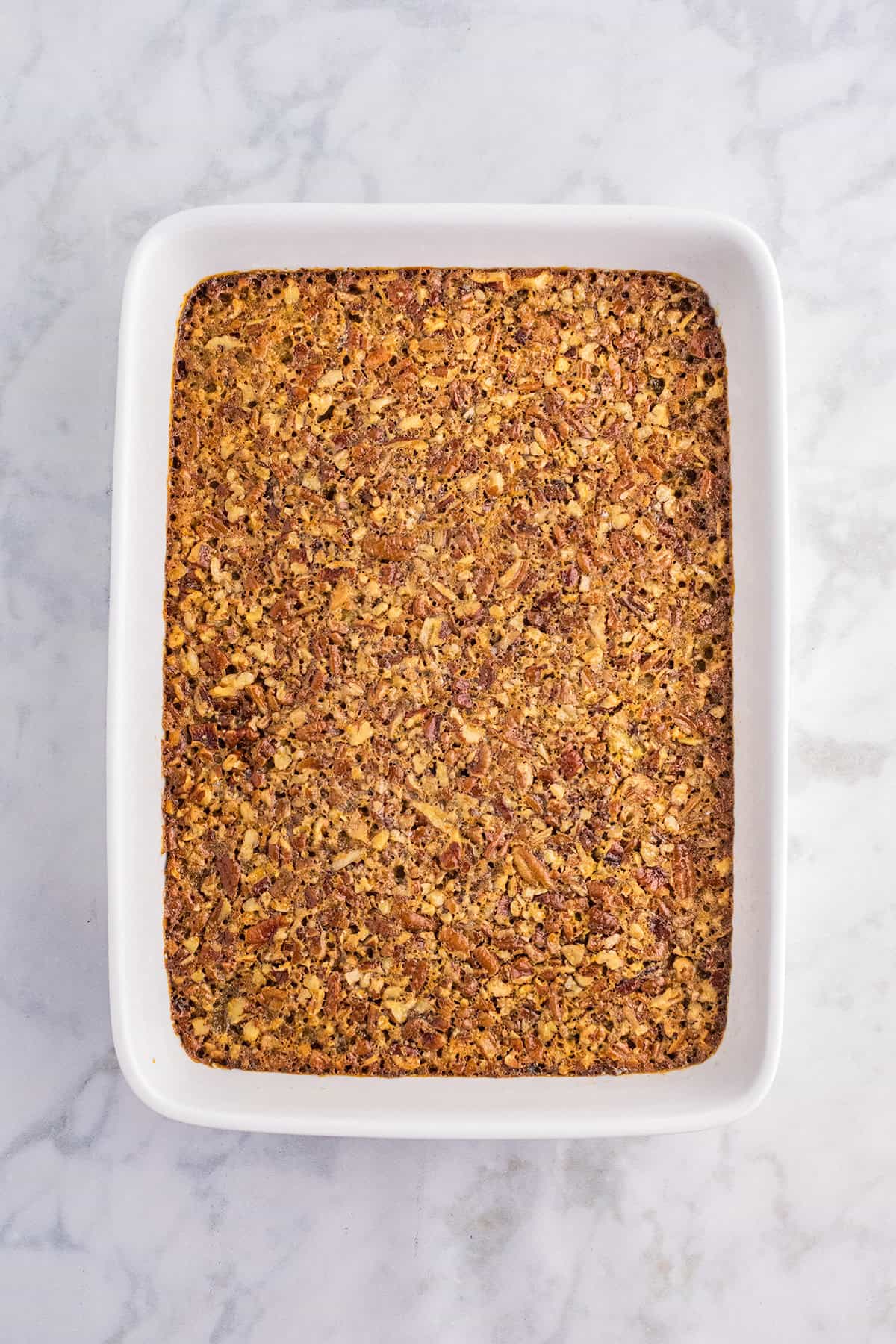 A rectangular baked casserole with a golden-brown, textured pecan pie bars-inspired nut topping in a white dish on a marble surface.