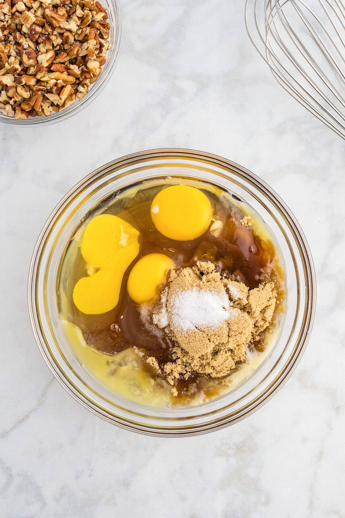 A glass bowl contains eggs, brown sugar, and a mound of white sugar on a marble surface, ready to be mixed for delicious Pecan Pie Bars. Nearby are chopped pecans in another bowl and a metal whisk.