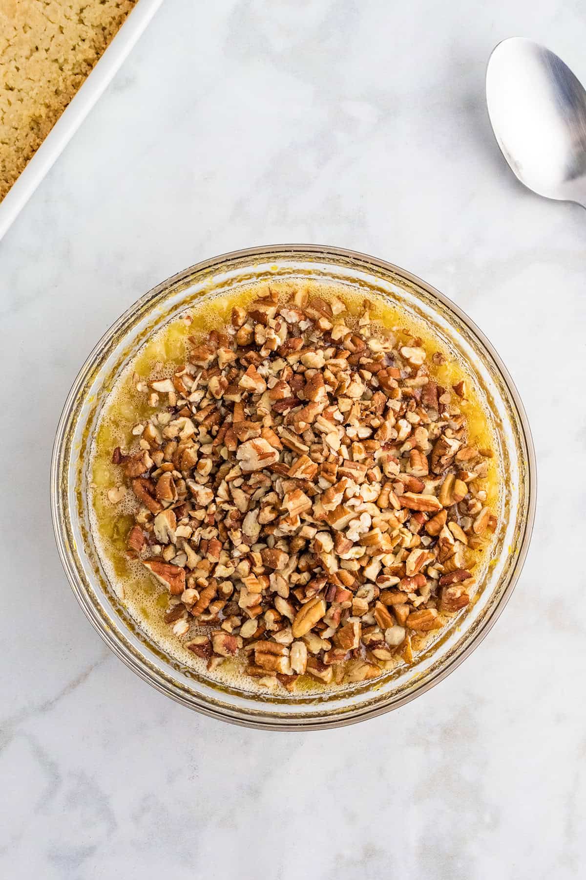 A glass bowl filled with melted butter and chopped pecans, perfect for making Pecan Pie Bars, sits on a marble surface next to a silver spoon and a corner of a baking dish.