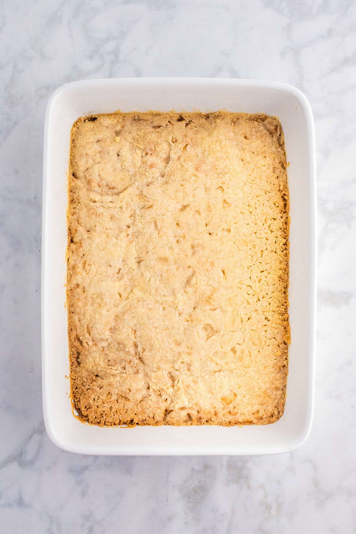A square, golden-brown baked dessert, resembling decadent Pecan Pie Bars, sits in a white rectangular ceramic dish on a marble surface.