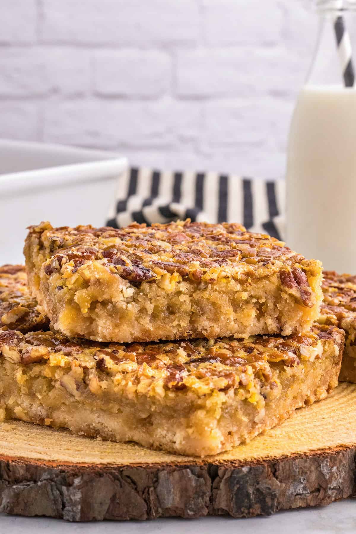 Two Pecan Pie Bars stacked on a wooden serving board, with a bottle of milk and a baking dish in the background.