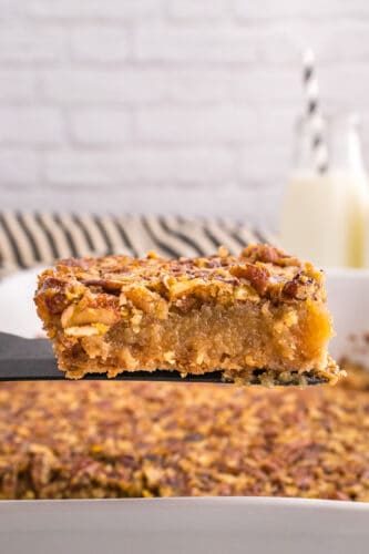 A close-up of a delicious Pecan Pie Bars dessert bar being lifted from a baking dish, with a glass of milk and striped straws in the background.