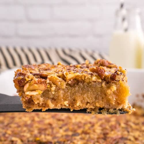 A close-up of a delicious Pecan Pie Bars dessert bar being lifted from a baking dish, with a glass of milk and striped straws in the background.