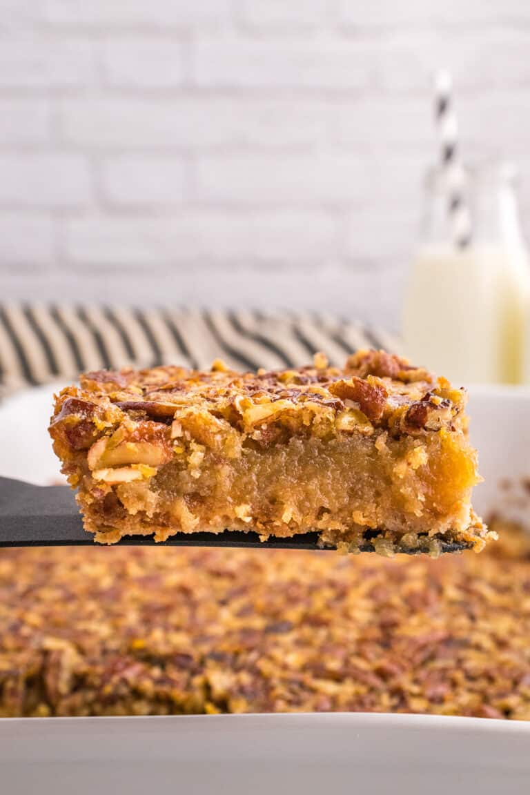 A close-up of a delicious Pecan Pie Bars dessert bar being lifted from a baking dish, with a glass of milk and striped straws in the background.