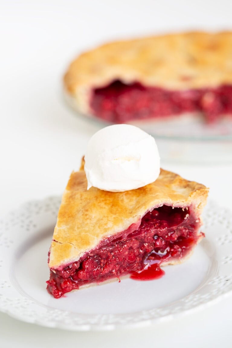 A slice of cherry and raspberry pie with a golden crust and a scoop of vanilla ice cream on top, served on a white plate, with the remaining pie in the background.