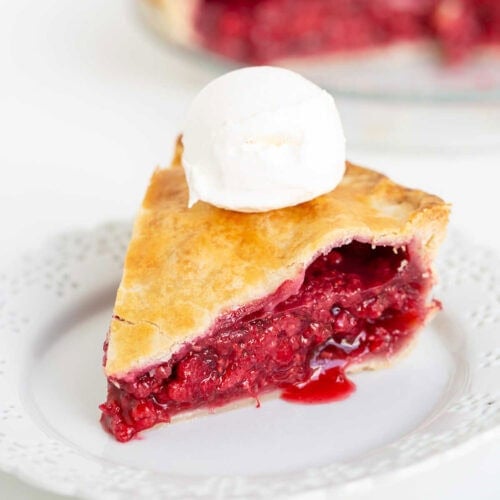 A slice of raspberry pie with a golden crust and a scoop of vanilla ice cream on top, served on a white plate.