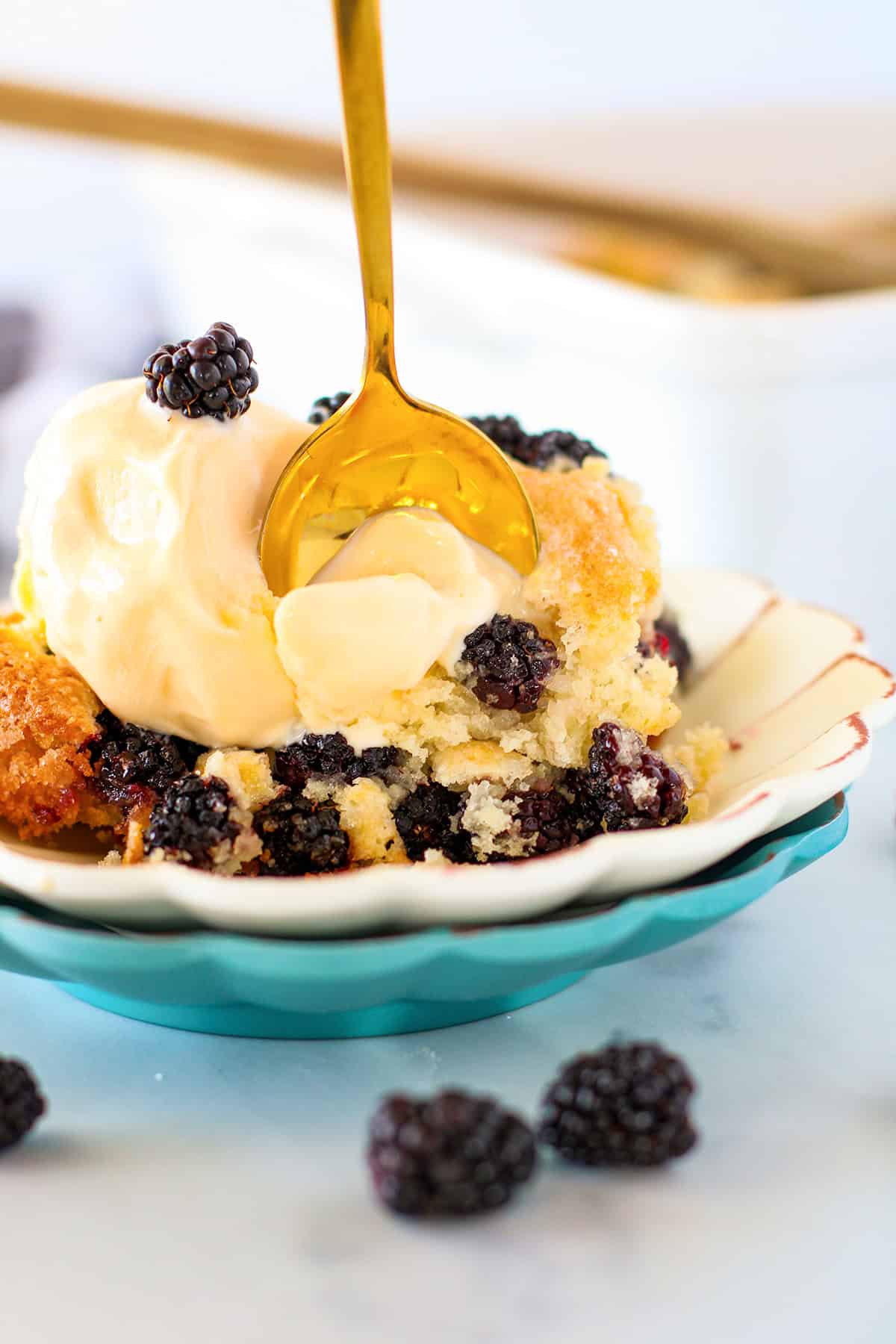 A serving of warm blackberry cobbler topped with vanilla ice cream is shown on a plate, with a gold spoon scooping into the luscious dessert. Fresh blackberries are scattered nearby, highlighting the rich flavors of this classic blackberry cobbler.
