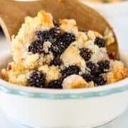 A wooden spoon serves a portion of blackberry cobbler from a white ceramic dish, revealing its golden crust and baked blackberries.