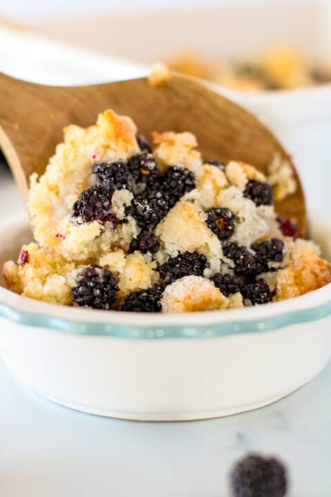 A wooden spoon serves a portion of blackberry cobbler from a white ceramic dish, revealing its golden crust and baked blackberries.