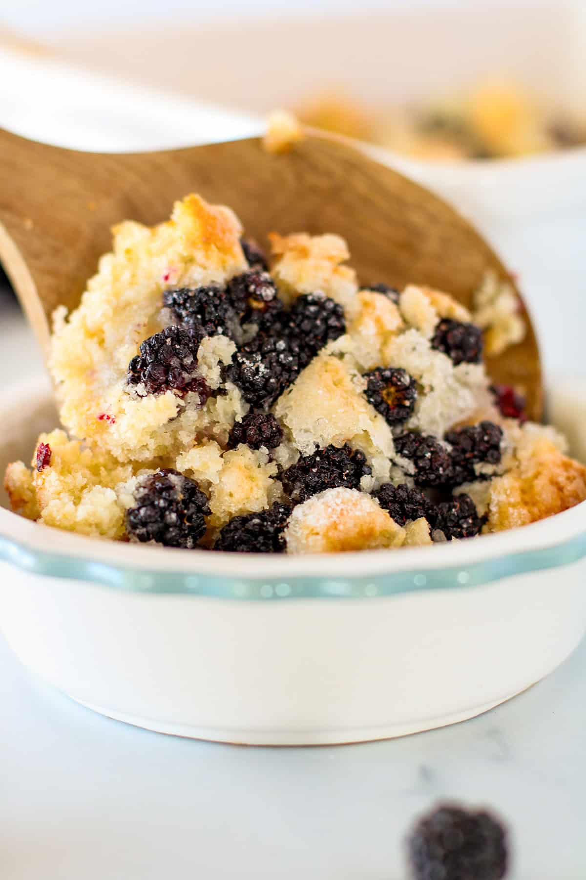 A wooden spoon serves a portion of blackberry cobbler from a white ceramic dish, revealing its golden crust and baked blackberries.
