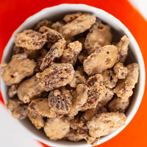 A white bowl filled with candied pecans sits on an orange napkin, showcasing the rich glaze and crunch of these irresistible candied pecans, viewed from above.